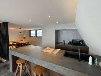 Modern kitchen with concrete countertops and wooden stools, open to a dining area with wooden table and chairs.