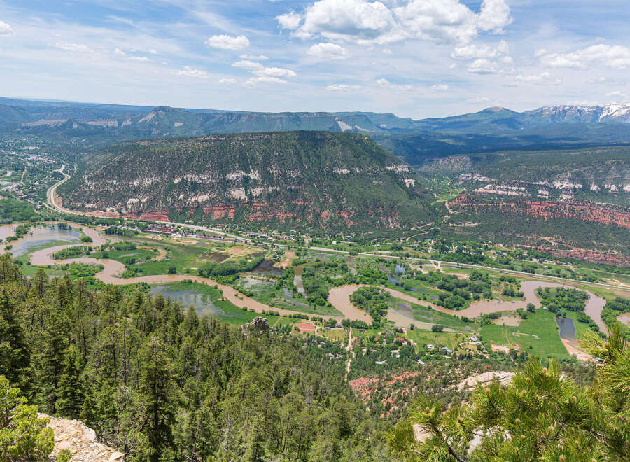Panoramic view of a lush green valley with a meandering river, surrounded by majestic mountains under a vibrant blue sky.