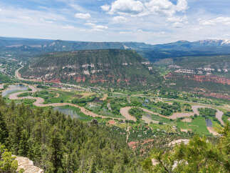 Panoramic view of a lush green valley with a meandering river, surrounded by majestic mountains under a vibrant blue sky.
