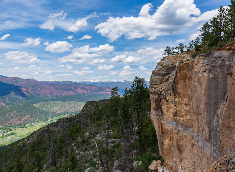 Spectacular vista of a mountainous landscape, showcasing a dramatic cliff edge with lush greenery and distant snow-capped peaks under a vibrant blue sky.