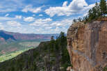 Spectacular vista of a mountainous landscape, showcasing a dramatic cliff edge with lush greenery and distant snow-capped peaks under a vibrant blue sky.