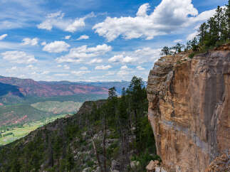 Spectacular vista of a mountainous landscape, showcasing a dramatic cliff edge with lush greenery and distant snow-capped peaks under a vibrant blue sky.