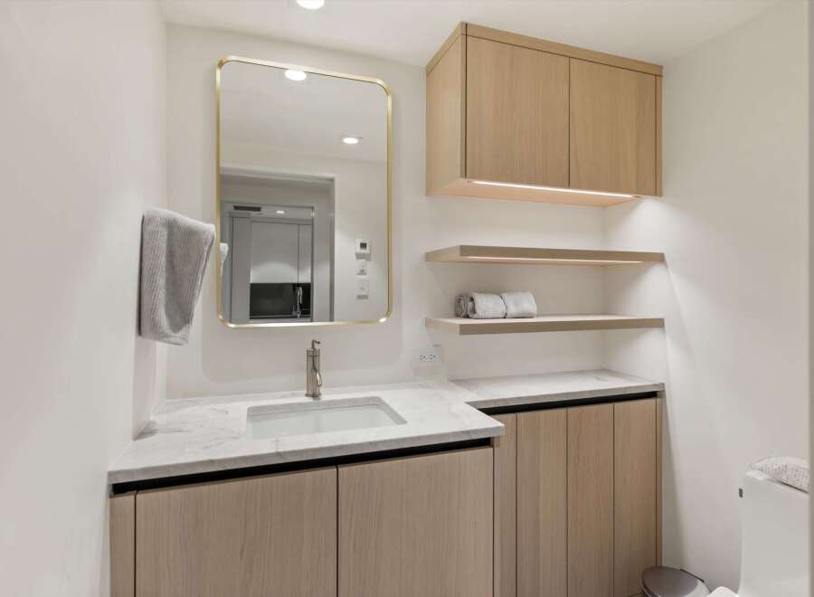 This stunning bathroom boasts a modern design with light oak wood cabinets, marble countertops, and a sleek gold-framed mirror.