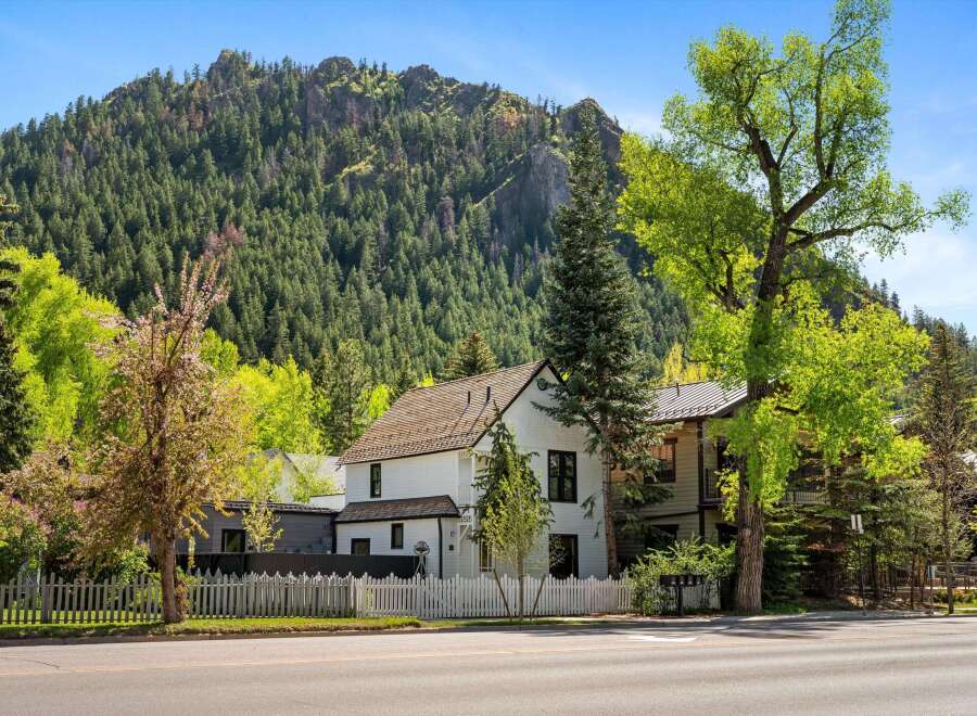 Charming white house nestled in a mountain setting, featuring a picket fence and mature trees.