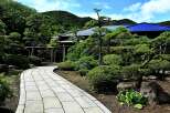 This serene Japanese garden features a winding stone pathway, meticulously manicured shrubs and trees, and a traditional house with a dark blue roof.