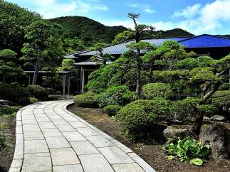 This serene Japanese garden features a winding stone pathway, meticulously manicured shrubs and trees, and a traditional house with a dark blue roof.