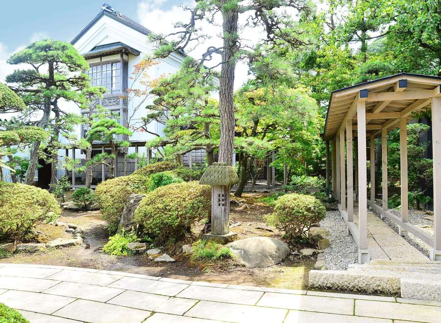 This serene Japanese garden features a traditional house with large windows, surrounded by meticulously manicured trees and shrubs.