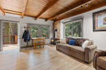 Cozy cabin living room with wood-beamed ceiling, wood flooring, stone fireplace, and large windows offering stunning forest views.
