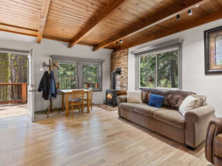 Cozy cabin living room with wood-beamed ceiling, wood flooring, stone fireplace, and large windows offering stunning forest views.