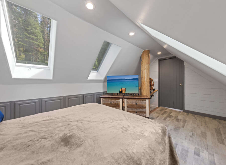 Cozy attic bedroom with plush bedding, rustic wood dresser, and a flat-screen TV showcasing a scenic lake view.