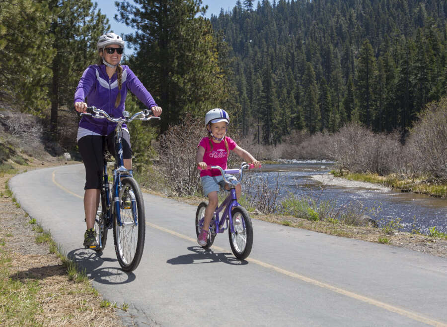 A mother and daughter enjoy a scenic bike ride along a paved path, with a tranquil river and lush forest as their backdrop.