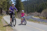 A mother and daughter enjoy a scenic bike ride along a paved path, with a tranquil river and lush forest as their backdrop.