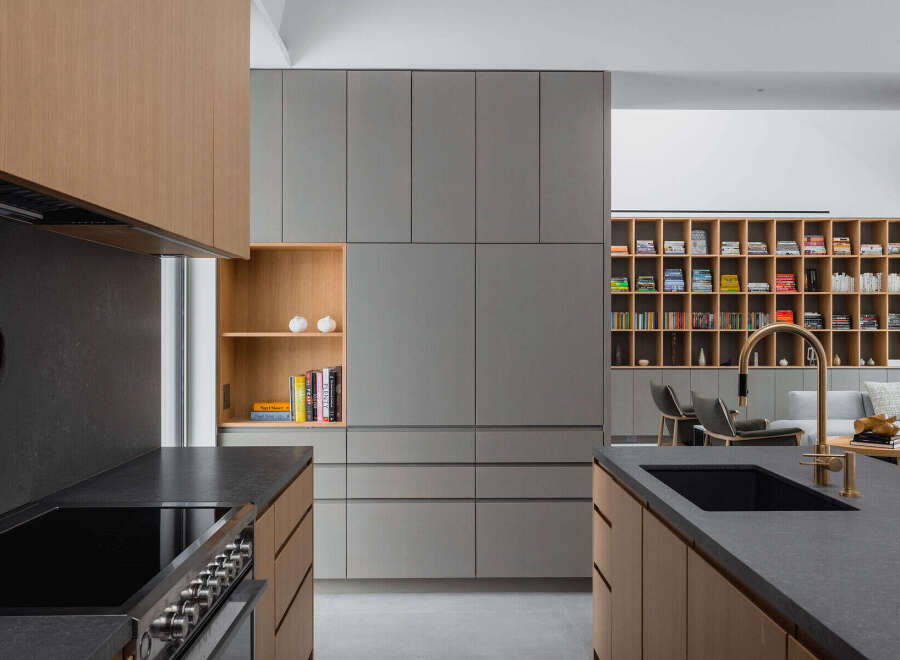 Modern kitchen with sleek gray and wood cabinetry, a built-in bookshelf, and a large island with a dark countertop and integrated sink.