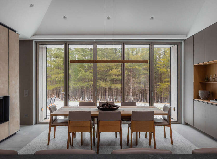 This stunning modern home boasts a spacious dining area with floor-to-ceiling windows offering breathtaking views of a snow-dusted forest.
