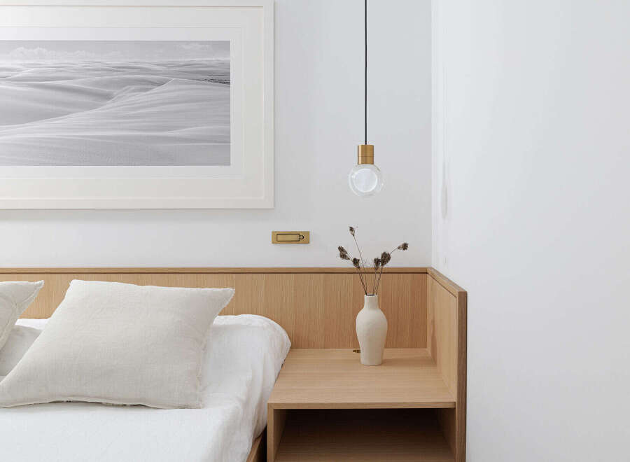 This serene bedroom boasts a minimalist aesthetic with a light oak wood headboard and nightstand, complemented by crisp white linens and a monochrome desert landscape print.