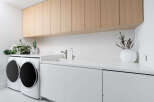 This laundry room boasts a clean, modern design with white cabinetry and countertops, complemented by light wood upper cabinets.