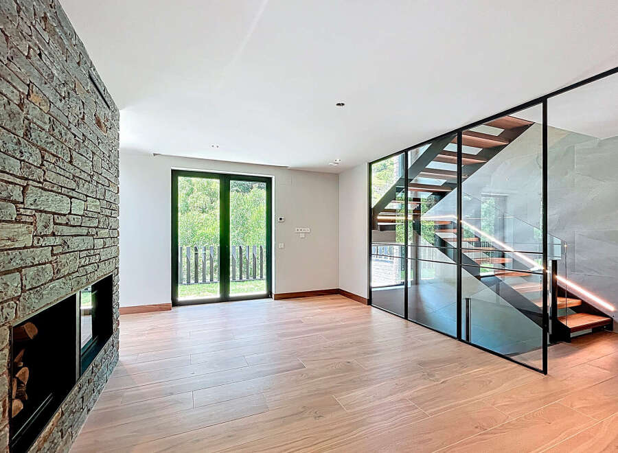 Discover this modern living space with a striking stone fireplace, a contemporary staircase enclosed in glass, and expansive outdoor views through large windows.