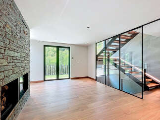 Discover this modern living space with a striking stone fireplace, a contemporary staircase enclosed in glass, and expansive outdoor views through large windows.