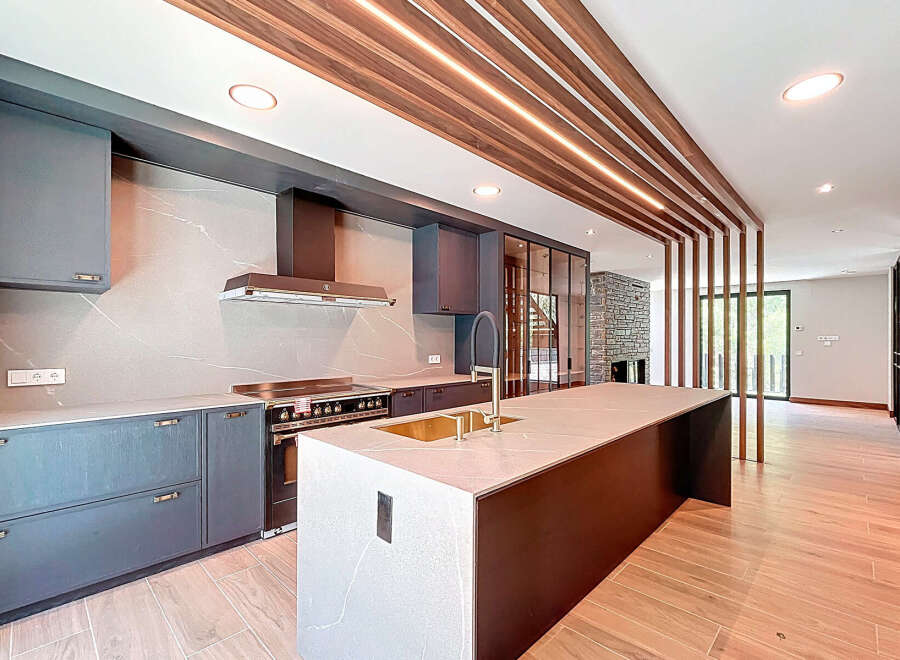 This modern kitchen features sleek gray cabinetry, a large central island with a gold sink, and a unique wooden slat ceiling design with integrated lighting.