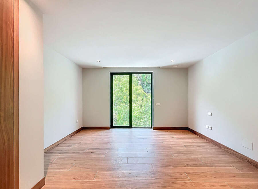 This bright, empty room features wood-look tile flooring and a large glass door offering views of lush greenery.