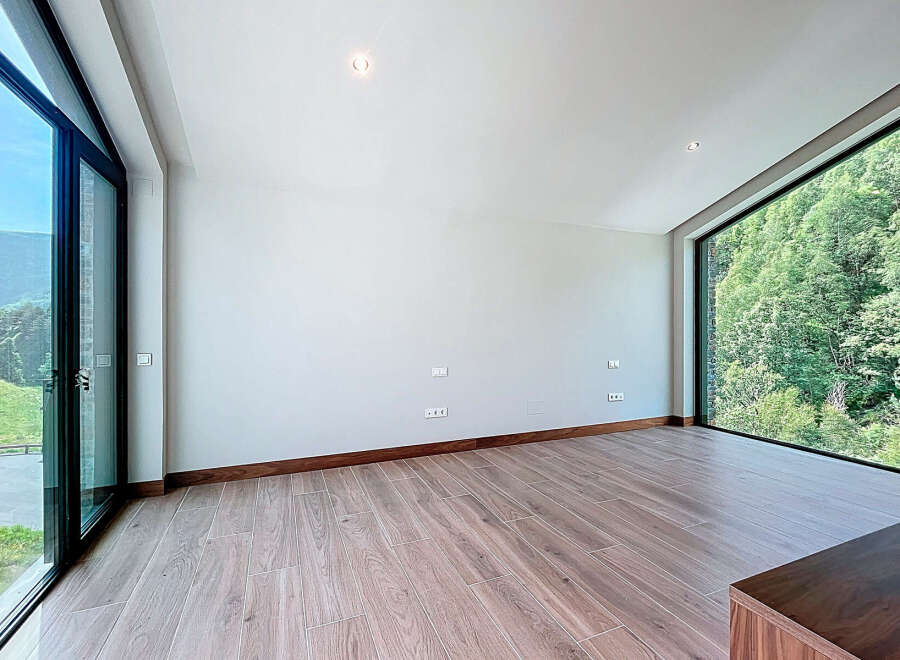 An expansive room features wood-look tile flooring and a natural wood baseboard, complemented by large windows that offer stunning views of the surrounding greenery.