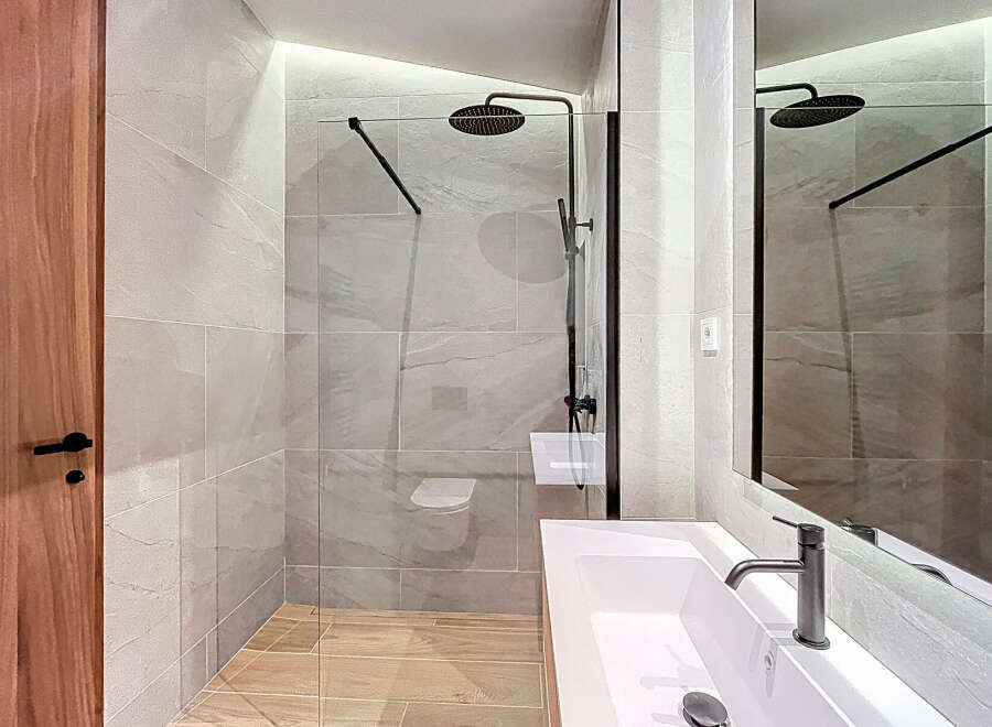 This modern bathroom features a sleek, frameless glass shower enclosure with a rainfall showerhead, complemented by light gray wall tiles and warm wood-look flooring.