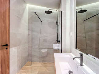 This modern bathroom features a sleek, frameless glass shower enclosure with a rainfall showerhead, complemented by light gray wall tiles and warm wood-look flooring.