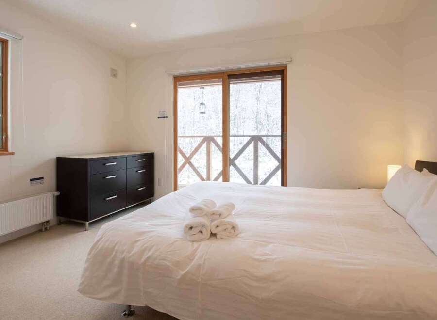 Experience cozy comfort in this inviting bedroom featuring a plush bed with fresh towels, a dark wood dresser for ample storage, and a window offering picturesque winter views.