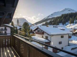 A scenic view from a wooden balcony showcases snow-covered buildings nestled in a mountain village.
