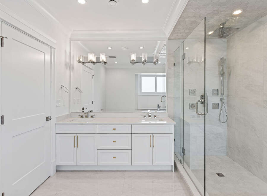 This pristine bathroom boasts a double vanity with white cabinets and gold hardware, complemented by a spacious glass-enclosed shower featuring marble-like tiling.