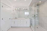 This pristine bathroom boasts a double vanity with white cabinets and gold hardware, complemented by a spacious glass-enclosed shower featuring marble-like tiling.