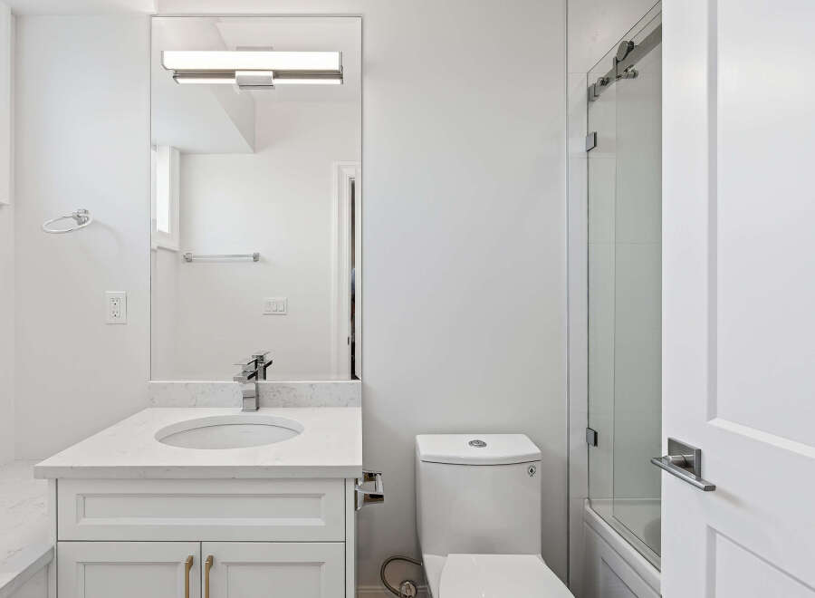 This is a well-lit, modern bathroom featuring a white vanity with a marble countertop, a sleek mirror with integrated lighting, and a toilet.