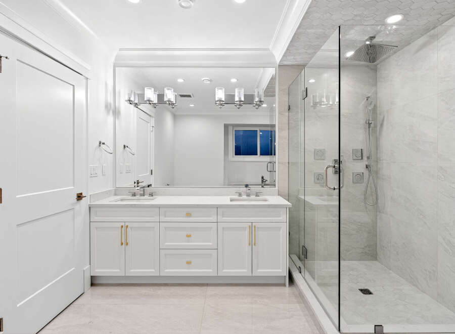 This is a bright and modern bathroom featuring a double vanity with gold hardware, a spacious walk-in shower with gray marble-style tiling and a rainfall showerhead.