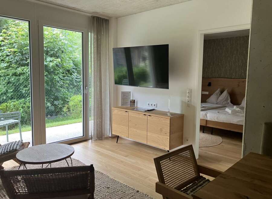 This property features a bright living area with a TV, a wooden cabinet, and large windows that offer a view of the green outdoors.