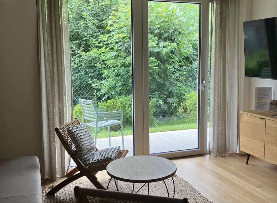 This inviting living space features stylish seating, a round coffee table, and a jute rug, all complemented by natural light streaming through large windows that offer views of lush greenery.