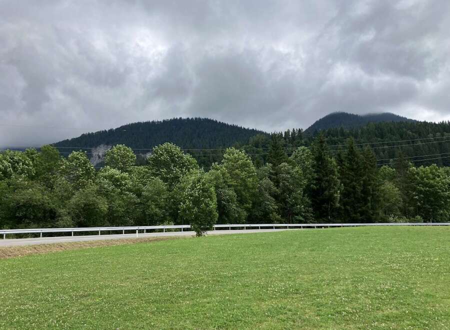 Discover a serene property with lush green fields, bordered by a quaint road and a backdrop of majestic mountains.
