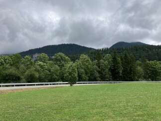 Discover a serene property with lush green fields, bordered by a quaint road and a backdrop of majestic mountains.