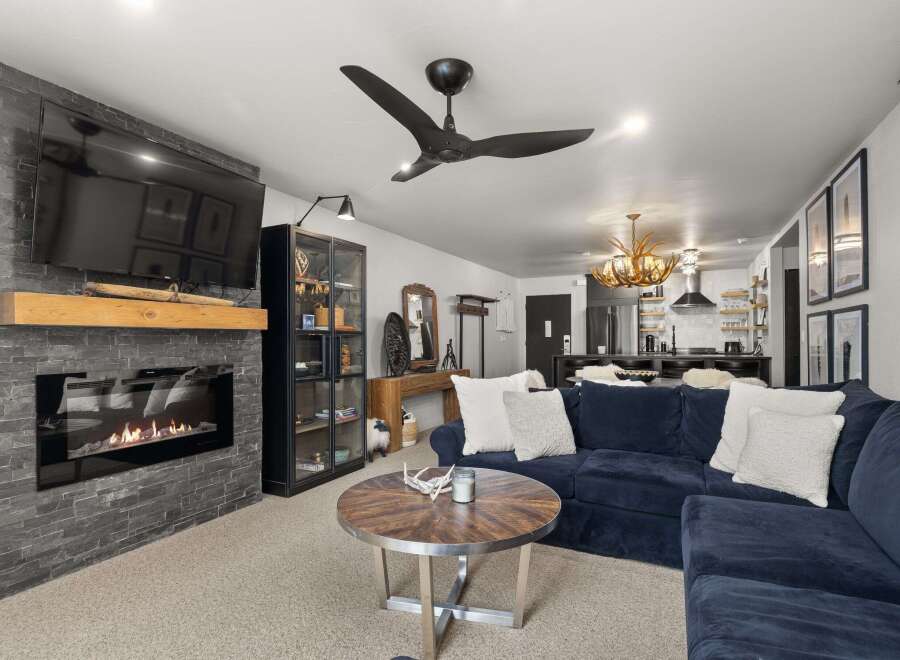 This inviting living room features a sleek fireplace with a stone surround and a rustic wooden mantel, complemented by a modern black ceiling fan.