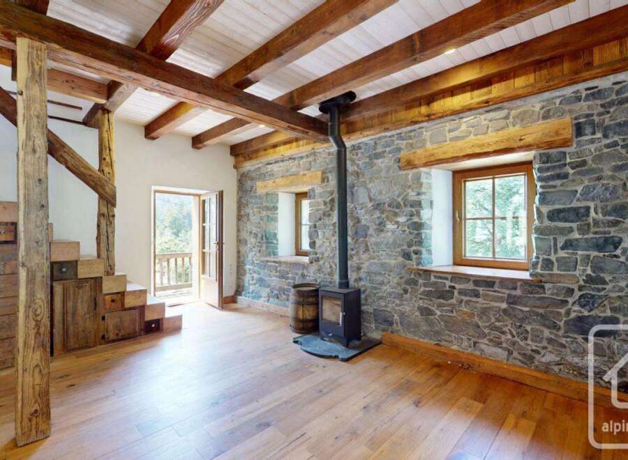 Discover this charming chalet featuring exposed wooden beams, a rustic stone wall, and a cozy wood-burning stove.
