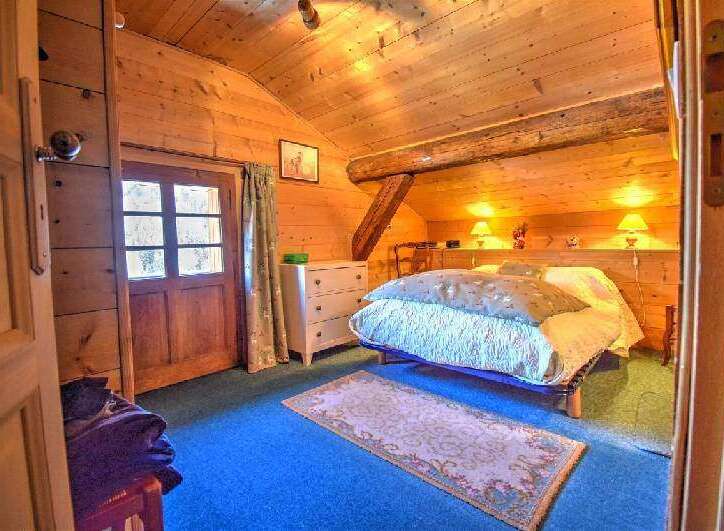 Discover this cozy wooden cabin bedroom featuring a sloped ceiling, a comfortable bed with bedside lamps, and a charming rug on a blue carpet.