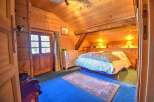 Discover this cozy wooden cabin bedroom featuring a sloped ceiling, a comfortable bed with bedside lamps, and a charming rug on a blue carpet.