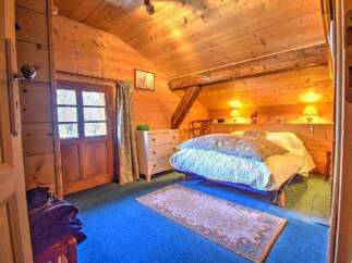 Discover this cozy wooden cabin bedroom featuring a sloped ceiling, a comfortable bed with bedside lamps, and a charming rug on a blue carpet.