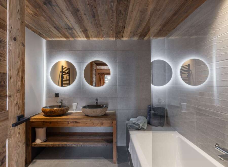 This bathroom features a rustic wooden vanity with stone vessel sinks, illuminated round mirrors, and a modern bathtub.