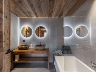 This bathroom features a rustic wooden vanity with stone vessel sinks, illuminated round mirrors, and a modern bathtub.