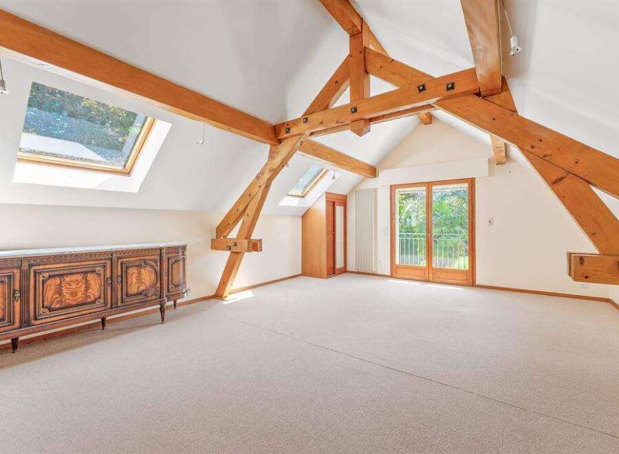 A spacious attic room featuring exposed wooden beams, skylights that flood the space with natural light, and elegant French doors opening to a balcony.