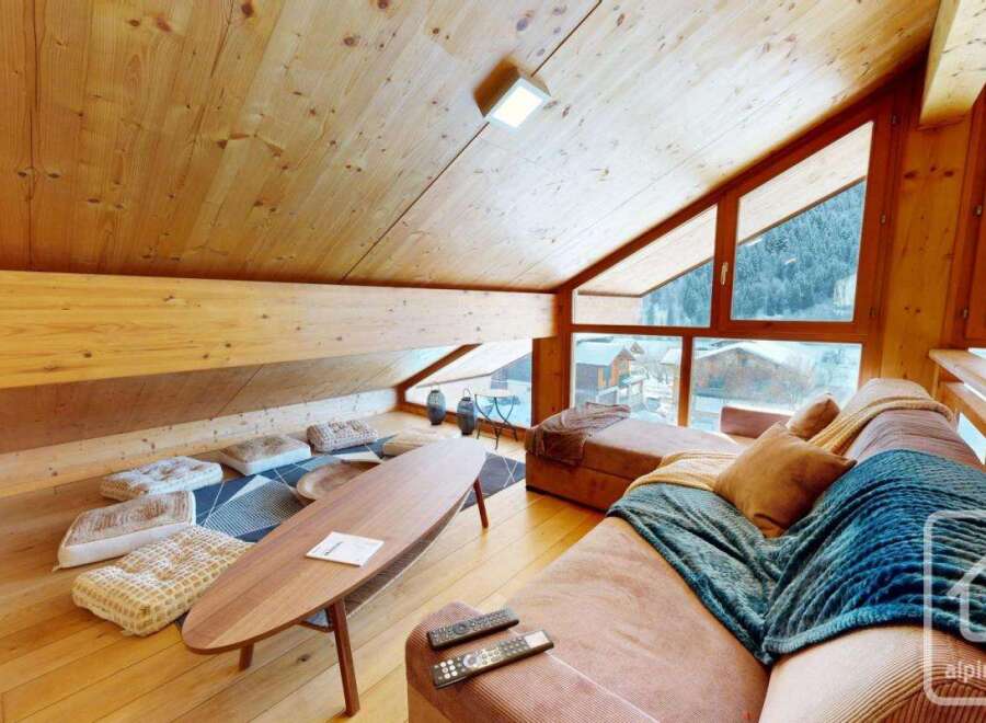 Experience the charm of this stunning alpine property, featuring a cozy living space with a wooden ceiling and large windows that offer breathtaking views of the surrounding mountains.