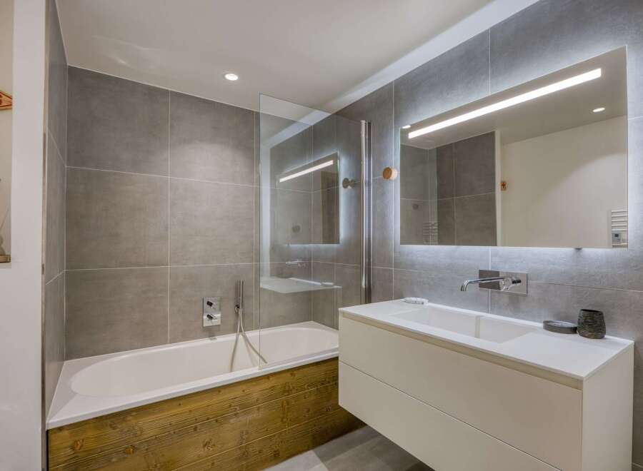 This modern bathroom features a sleek design with gray tiled walls and a wooden bathtub surround.