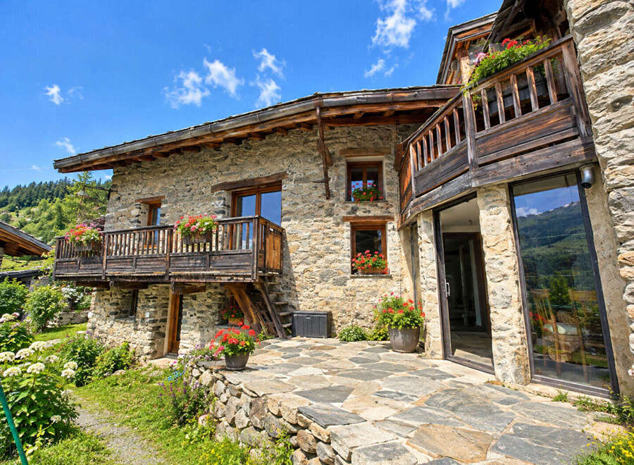 Experience rustic charm in this stone mountain home, featuring wooden balconies adorned with vibrant flowers and expansive windows offering breathtaking views.