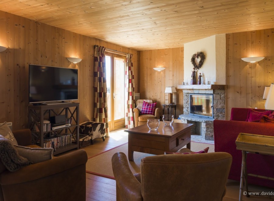 Cozy chalet living room with wooden walls and beamed ceiling, featuring a fireplace, comfortable leather armchairs, and a large flat-screen TV.