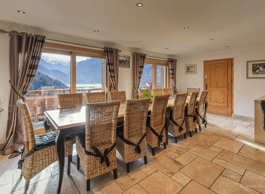 This stunning chalet boasts a spacious dining area with a large table perfect for family gatherings, surrounded by wicker chairs and offering breathtaking mountain views through expansive windows.